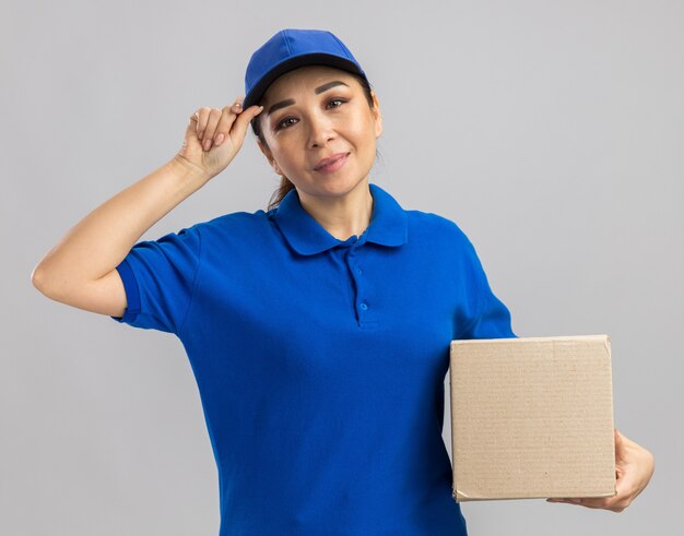 Jovem feliz entregadora de uniforme azul e boné segurando uma caixa de papelão com um sorriso no rosto em pé sobre uma parede branca