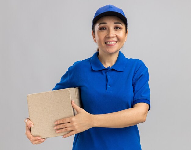 Jovem feliz entregadora de uniforme azul e boné segurando uma caixa de papelão com um sorriso no rosto em pé sobre uma parede branca