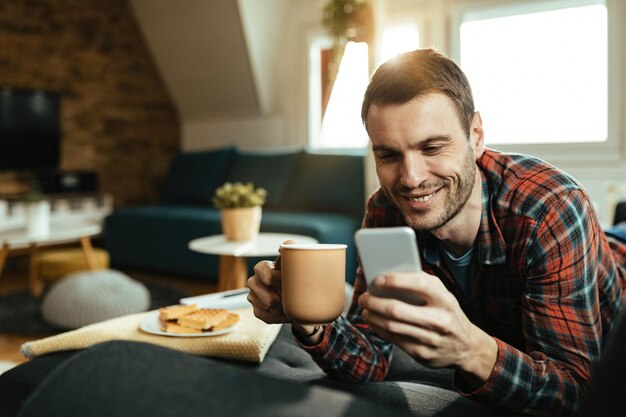 Jovem feliz lendo mensagem de texto no telefone inteligente enquanto bebe café e relaxa na sala de estar