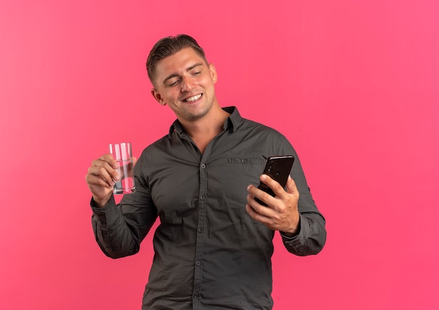 Jovem feliz loira bonito segurando um copo de água e olhando para o telefone isolado em um fundo rosa com espaço de cópia