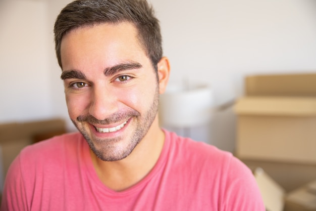 Jovem feliz se mudando para um novo apartamento, parado em frente a uma pilha de caixas de papelão abertas, olhando para a câmera