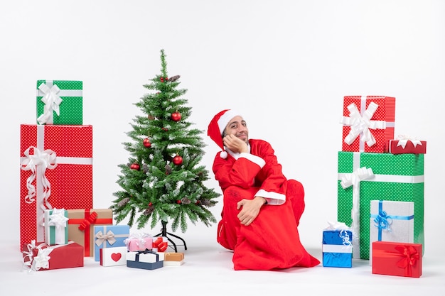 Jovem feliz sorridente vestido de papai noel com presentes e uma árvore de natal decorada sentada no chão sobre fundo branco