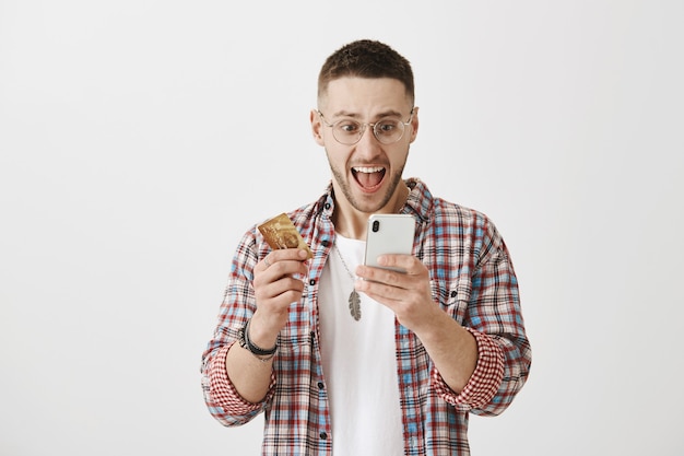 Jovem feliz surpreso com óculos posando com seu telefone e cartão