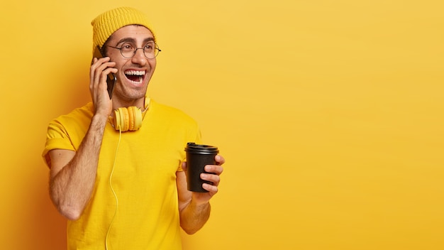 Jovem feliz tem uma boa conversa pelo celular, segura um café para viagem, gosta de beber, vestido com uma camiseta casual e um chapéu