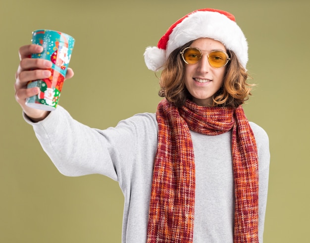 Jovem feliz usando chapéu de papai noel de natal e óculos amarelos com lenço quente em volta do pescoço mostrando copo de papel colorido sorrindo alegremente em pé sobre fundo verde