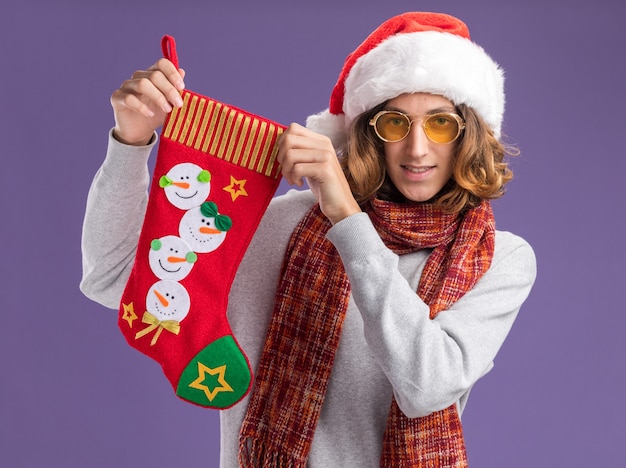 Foto grátis jovem feliz usando chapéu de papai noel de natal e óculos amarelos com um lenço quente no pescoço segurando uma meia de natal, olhando para a câmera com um sorriso no rosto em pé sobre um fundo roxo