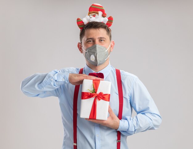 Jovem feliz usando suspensórios gravata borboleta na borda com Papai Noel usando máscara facial protetora segurando um presente, olhando para a câmera em pé sobre um fundo branco