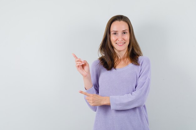 Jovem fêmea apontando para cima enquanto sorri com uma blusa lilás e parecendo feliz