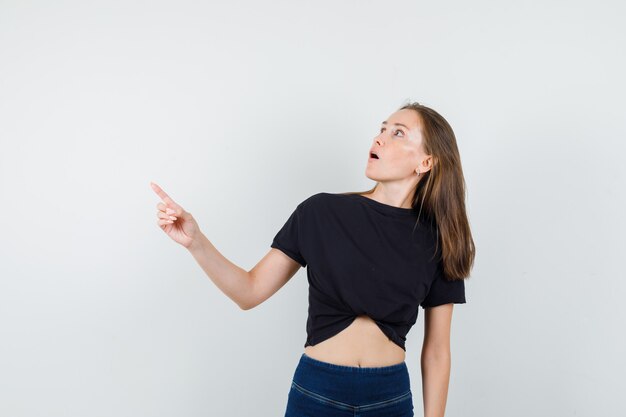 Jovem fêmea apontando para longe em blusa preta, calça e parecendo espantada,