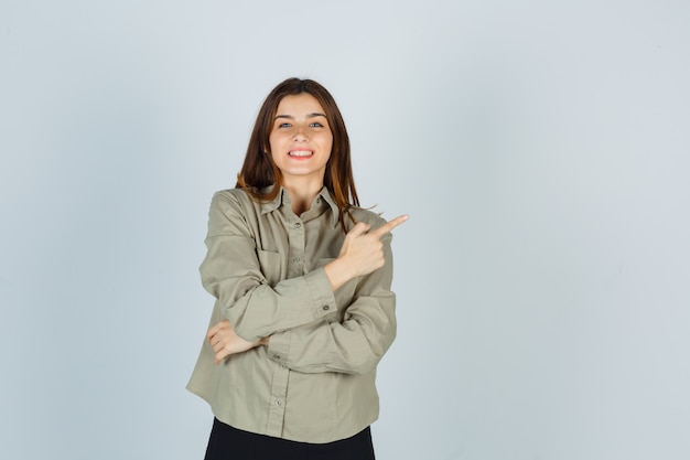 Jovem fêmea apontando para o canto superior direito da camisa, saia e parecendo feliz