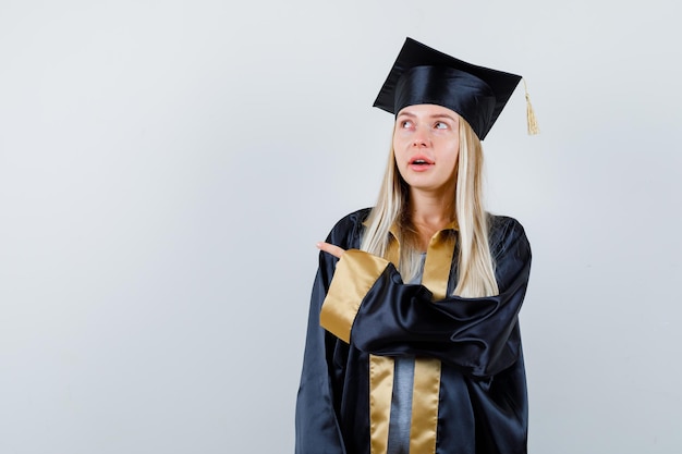 Jovem fêmea apontando para o lado em uniforme de pós-graduação e olhando pensativa.