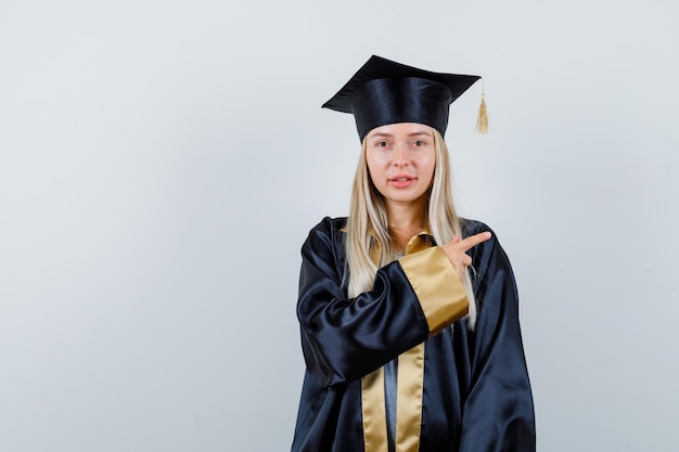 Jovem fêmea apontando para o lado em uniforme de pós-graduação e parecendo confiante.