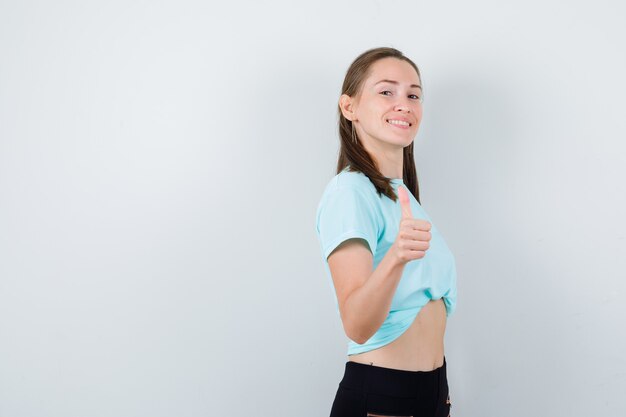 Jovem fêmea bonita aparecendo o polegar em t-shirt e olhando alegre, vista frontal.