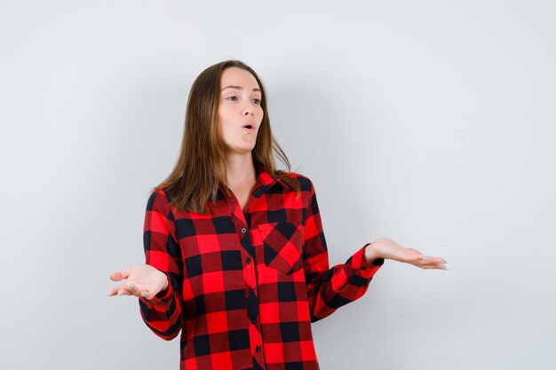 Jovem fêmea bonita mostrando um gesto impotente, olhando para longe na camisa casual e parecendo confusa. vista frontal.