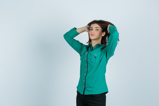 Jovem fêmea com camisa verde, calças posando com as mãos no cabelo castanho e olhando atraente, vista frontal.