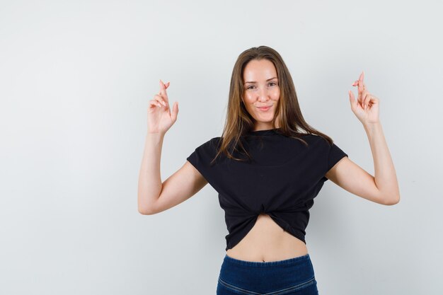 Jovem fêmea com os dedos cruzados na blusa e calça pretas e parecendo esperançosa