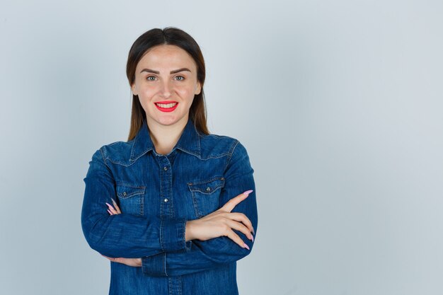 Jovem fêmea em pé com os braços cruzados em uma camisa jeans e parecendo feliz