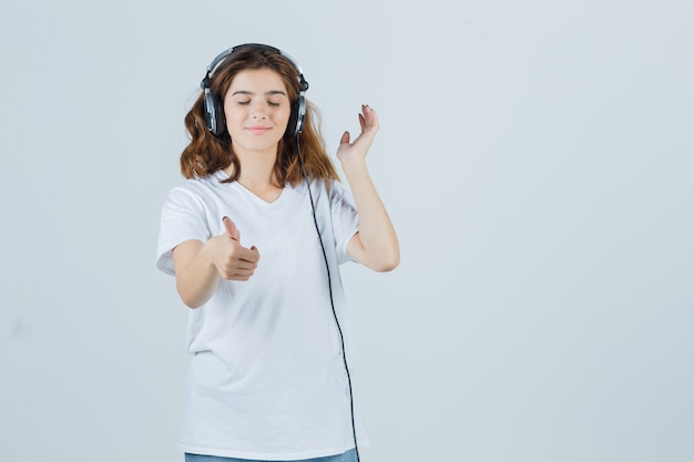 Jovem fêmea em t-shirt branca, jeans aparecendo o polegar enquanto ouve música com fones de ouvido e parece feliz, vista frontal.