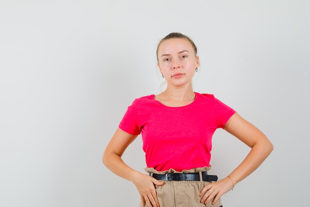 Jovem fêmea em t-shirt, calças posando em pé e olhando atraente, vista frontal.