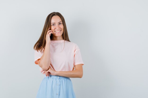 Jovem fêmea em t-shirt, saia falando no celular e olhando alegre, vista frontal.