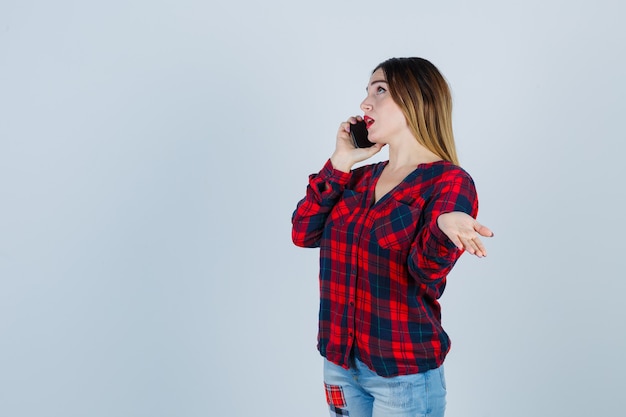 Jovem fêmea linda em camisa casual, falando no telefone, espalhando a palma da mão de lado e olhando pensativa, vista frontal.