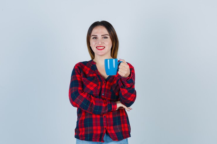 Foto grátis jovem fêmea linda em camisa casual, jeans, segurando a xícara enquanto posava e olhando alegre, vista frontal.