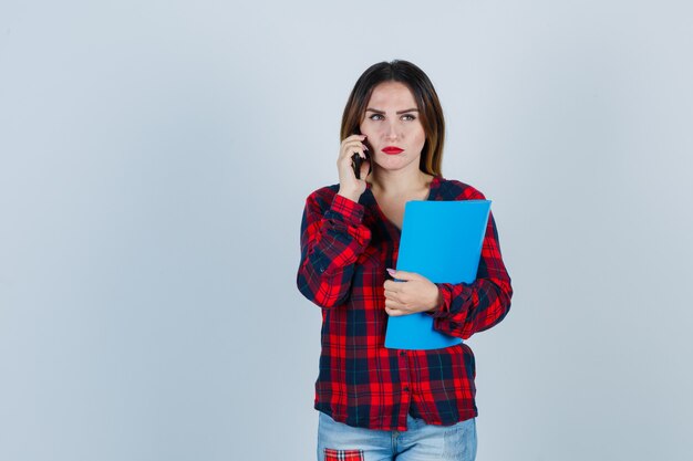 Jovem fêmea linda em camisa casual, jeans segurando uma pasta enquanto falava no telefone, olhando para longe e olhando mal-humorada, vista frontal.
