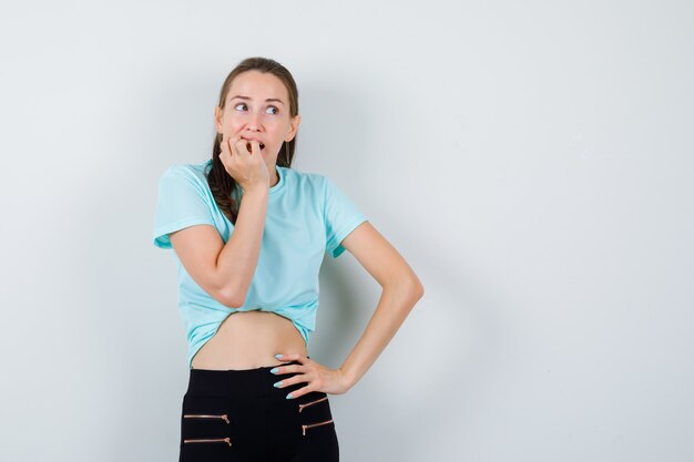 Jovem fêmea linda roendo as unhas, olhando para longe em camiseta, calça e parecendo estressada. vista frontal.