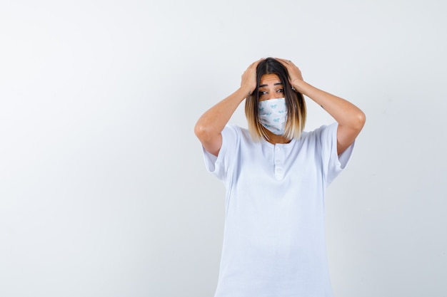 Jovem fêmea mantendo as mãos na cabeça em t-shirt, máscara e parecendo esquecido. vista frontal.