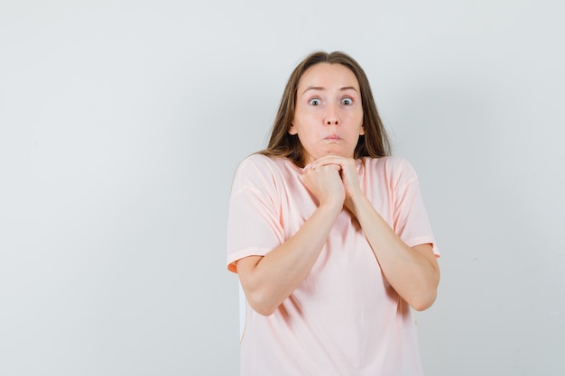 Foto grátis jovem fêmea mantendo as mãos postas sob o queixo em t-shirt rosa e olhando perplexa, vista frontal.