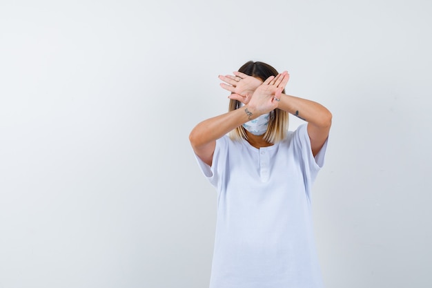 Jovem fêmea mostrando gesto de parada em t-shirt, máscara e olhando sério, vista frontal.