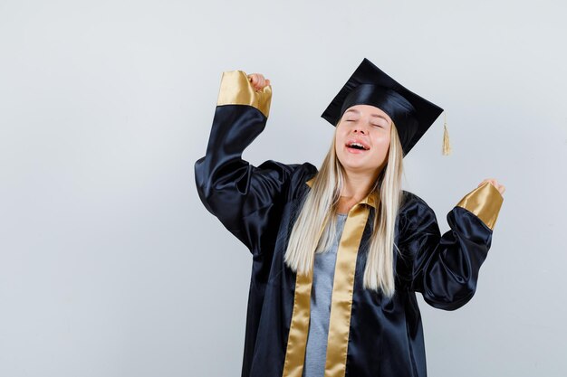 Jovem fêmea mostrando gesto de vencedor em uniforme de pós-graduação e parecendo feliz.