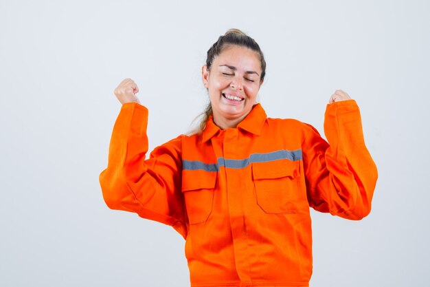 Jovem fêmea mostrando gesto de vencedor em uniforme de trabalhador e olhando alegre, vista frontal.