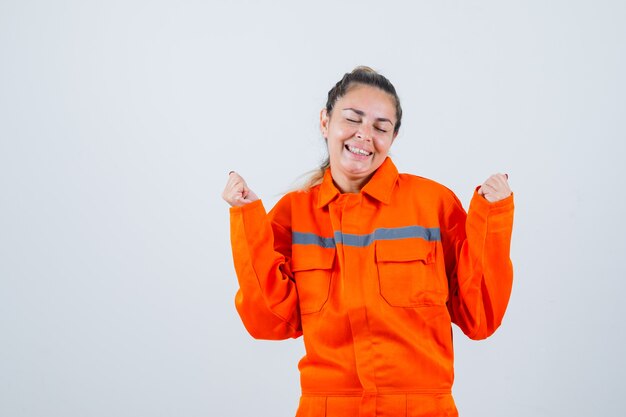 Jovem fêmea mostrando gesto de vencedor em uniforme de trabalhador e olhando alegre, vista frontal.