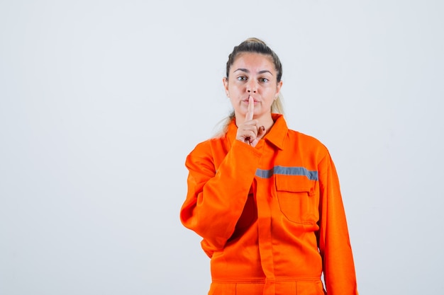 Jovem fêmea mostrando gesto silencioso em uniforme de trabalhador e olhando concentrada. vista frontal.