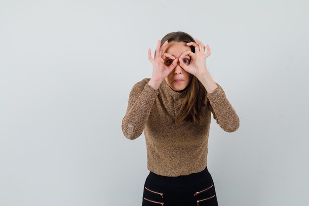 Foto grátis jovem fêmea na blusa dourada, mostrando o gesto binocular e olhando a vista frontal, focada. espaço para texto