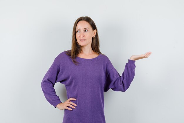 Jovem fêmea na camisa violeta, levantando a palma da mão como segurando algo e olhando pensativa, vista frontal.