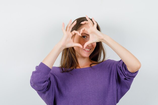 Jovem fêmea na camisa violeta, mostrando o gesto do coração e olhando alegre, vista frontal.