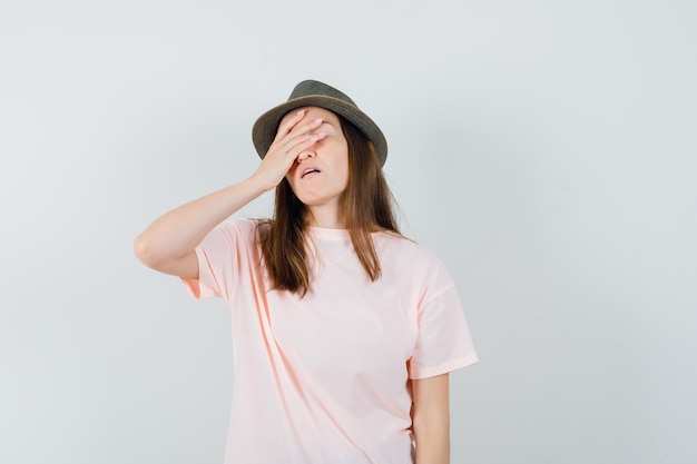Jovem fêmea segurando a mão no rosto em t-shirt rosa, chapéu e parecendo esquecido. vista frontal.