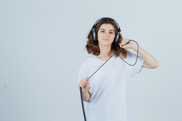Jovem fêmea segurando fones de ouvido em t-shirt branca e olhando alegre, vista frontal.