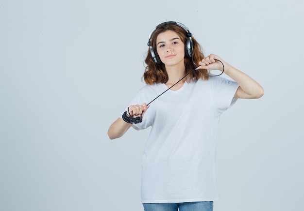 Jovem fêmea segurando fones de ouvido em t-shirt branca e olhando alegre, vista frontal.