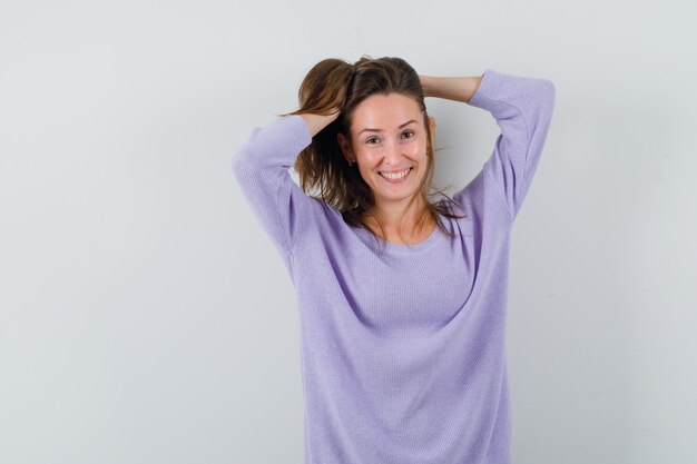 Jovem fêmea segurando o cabelo com as mãos na blusa lilás e parecendo alegre. vista frontal.