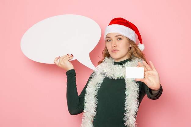 Jovem fêmea segurando uma grande placa branca e um cartão do banco no modelo de parede rosa, férias natal