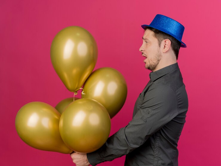 Foto grátis jovem festeiro impressionado com chapéu azul segurando e olhando para balões isolados em rosa