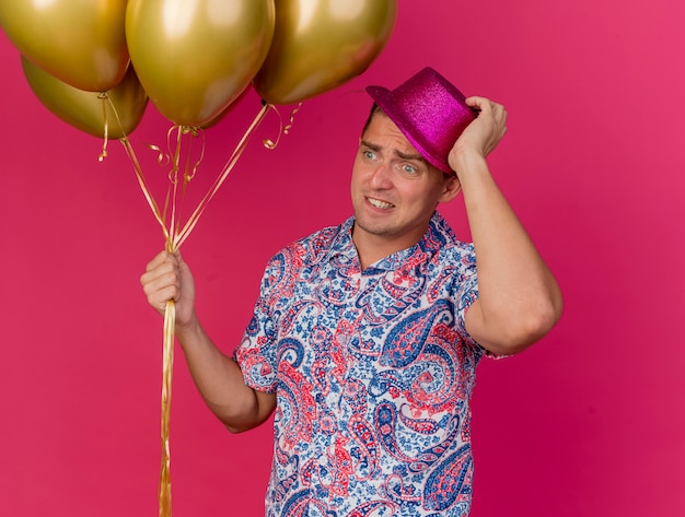 Jovem festeiro preocupado, olhando para o lado usando um chapéu rosa, segurando balões e segurando um chapéu isolado no fundo rosa