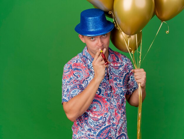 Jovem festeiro satisfeito com um chapéu azul, segurando balões e soprando um soprador de festa isolado no verde