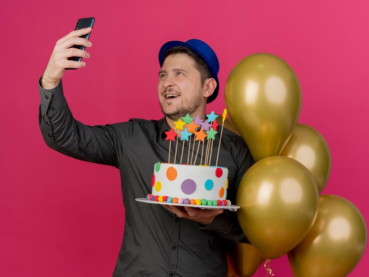 Foto grátis jovem festeiro sorridente com chapéu azul parado perto de balões segurando um bolo e tirando uma selfie isolada na rosa