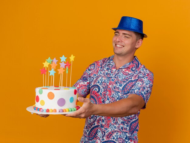 Jovem festeiro sorridente com chapéu azul segurando um bolo ao lado isolado em laranja