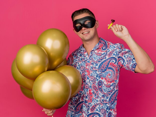 Jovem festeiro sorridente usando máscara de máscara segurando balões com soprador de festa isolado em rosa