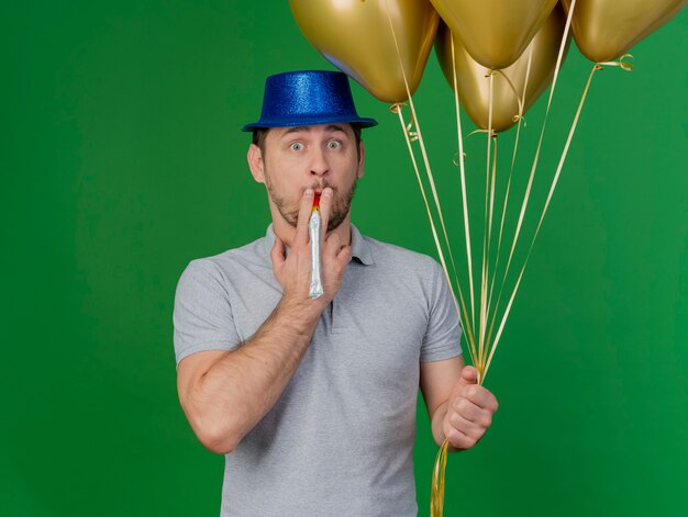 Jovem festeiro surpreso com chapéu de festa, apitando e segurando balões isolados no verde
